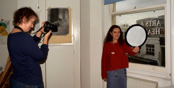 A young lady in a red jumper is standing with her left side by a window holding a small reflector in front of her while an older lady in a blue top is taking portrait photos of her. Photo by Eva Kalpadaki.