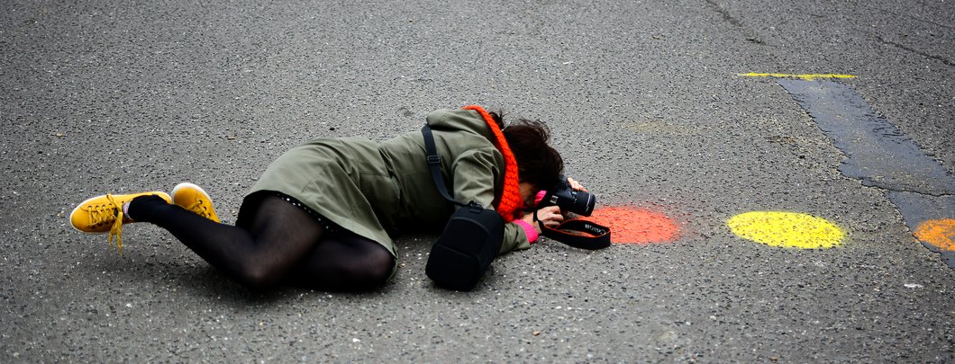 Female Bright-On-Photography student with yellow shoes and red scarf is lying down on the cement in Hove seafront and taking photographs  of some red and yellow painted dots. Photo by Eva Kalpadaki.