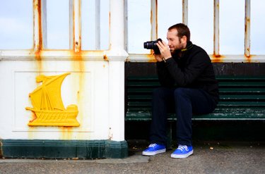 Male student is shown sitting on a Hove seafront shed taking photographs while holding his camera in some distance from his eyes. A yellow ship emblem appears on the side of the shed. Photo by Eva Kalpadaki.