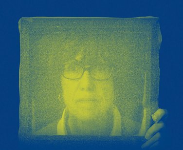 Portrait photo from above the shoulders of a woman wearing glasses through a yellow mouslin which she is holding with her left hand in a blue background border. Photo by Sue Daly.