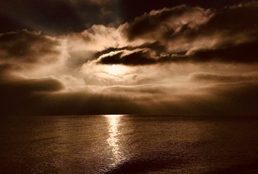Sepia photo showing sun behind dramatic clouds while it is reflected on the sea. Photo by Eva Kalpadaki.