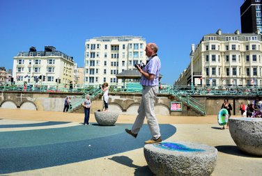 A Bright-on-photography male student is making a step in the air while stepping down from one of the half-stone eggs in Brighton seafront while three buildings are seen in the background. Photo by Eva Kalpadaki.